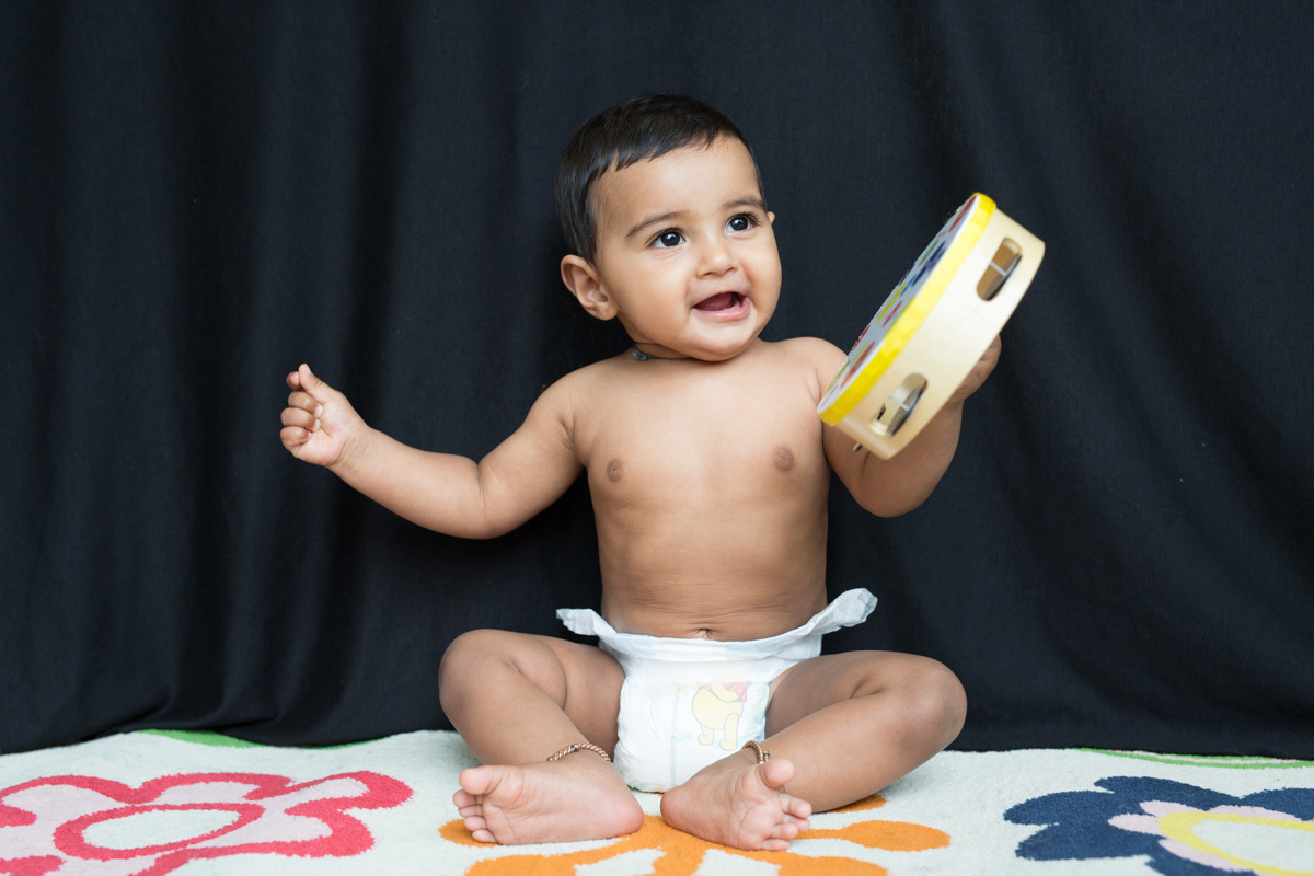 A 12 month old baby sits in front of a black curtain on a white rug with bright coloured flowers. The baby is holding a tambourine